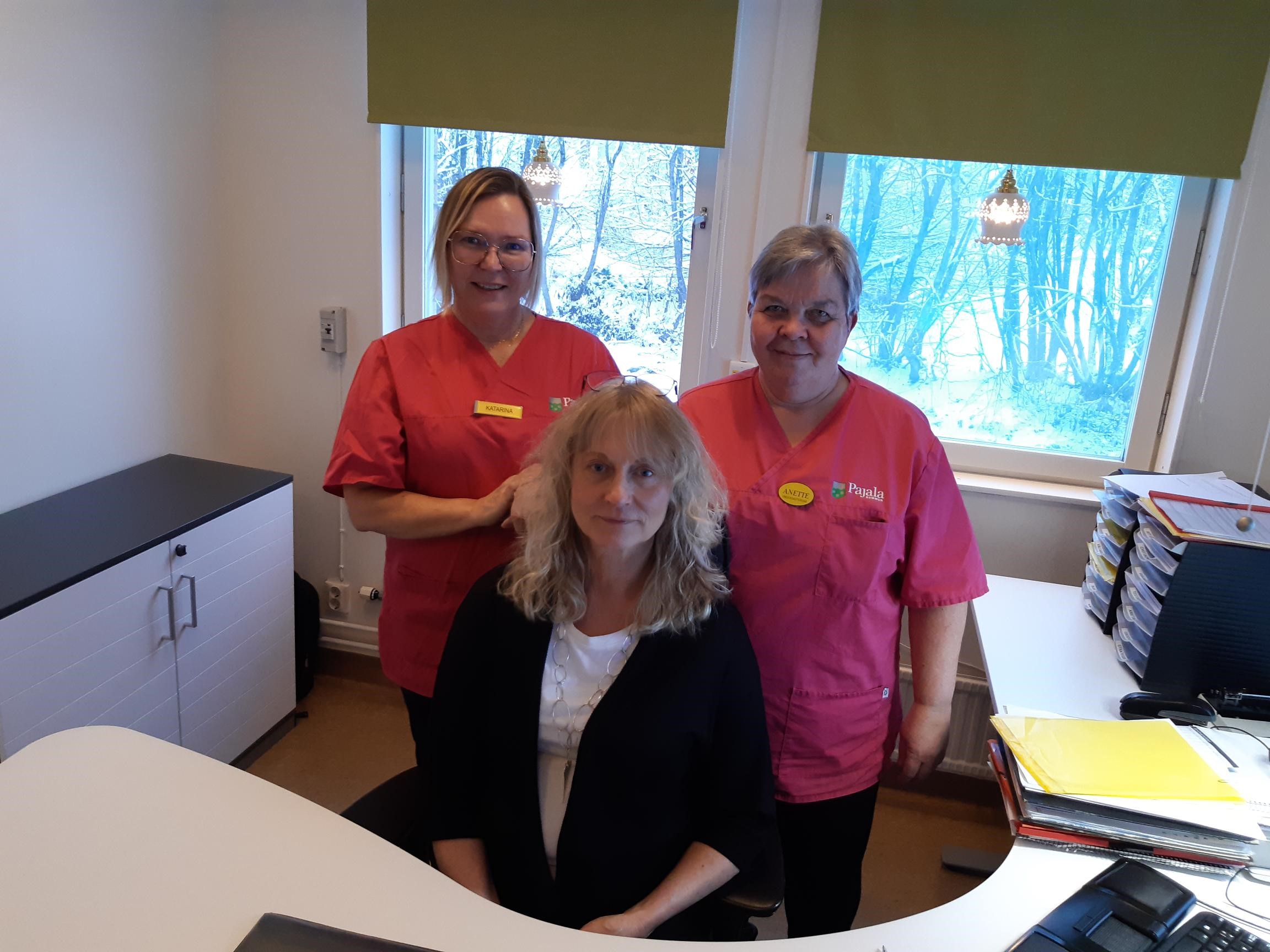 Fotot taget i ett kontor, på stol i mitten sitter Birgitta Uusitalo. Bakom henne står Katarina Henriksson och Anette Unga i sina arbetskläder. De har rosa tröjor och svarta byxor. 